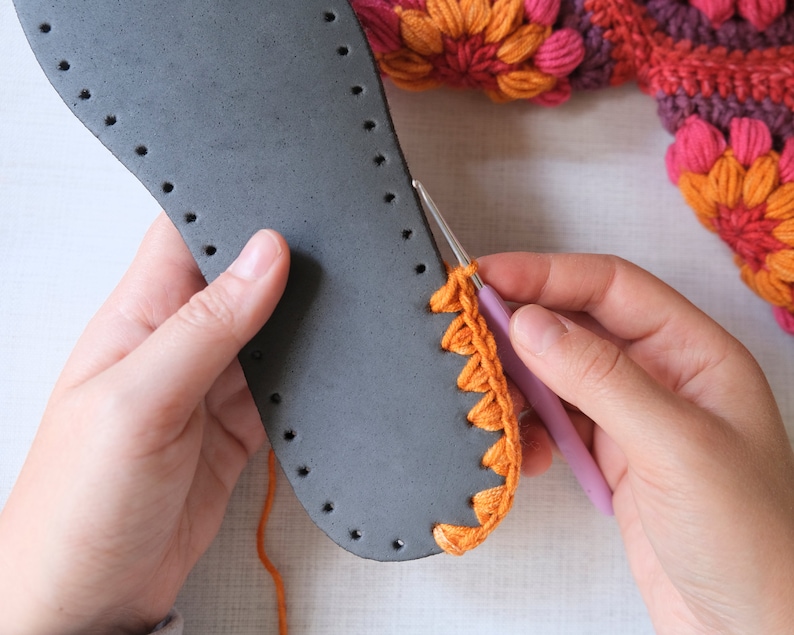 May include: A person is crocheting a bright orange crocheted edge onto a gray leather sole. The crocheted edge is being attached to the sole with a pink crochet hook.