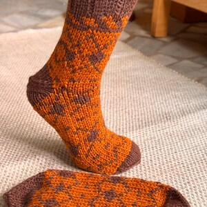 Puede incluir: Calcetines tejidos a mano en naranja y marrón con un patrón geométrico. Los calcetines tienen un puño y talón acanalados. Un calcetín se usa, el otro está plano. Hecho de hilo grueso.