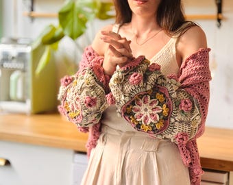 Puede incluir: Un cárdigan de ganchillo con una base de color rosa empolvado y motivos florales de cuadrados de la abuela en las mangas. Las mangas incorporan adornos de rosas en 3D con texturas en tonos rosa, crema y amarillo, sobre una camiseta sin mangas crema.