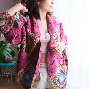 Puede incluir: Una chaqueta de punto de ganchillo rosa con patrones geométricos coloridos. La chaqueta está abierta por delante y tiene un corte holgado.