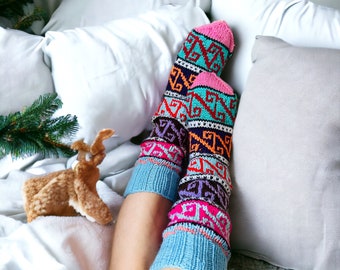 May include: A pair of colorful knit socks with a geometric pattern in shades of pink, teal, orange, and purple. The socks are on a bed with white pillows and a decorative reindeer.