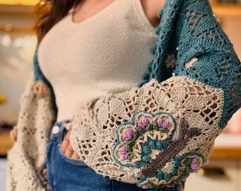 May include: A handmade crochet cardigan with a butterfly design on the sleeve. The cardigan features a teal and beige color scheme, with intricate openwork patterns. The sleeves have a beige butterfly design with pink and teal accents.