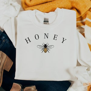 May include: White sweatshirt with the word "HONEY" printed in black letters and a black and yellow bee illustration.