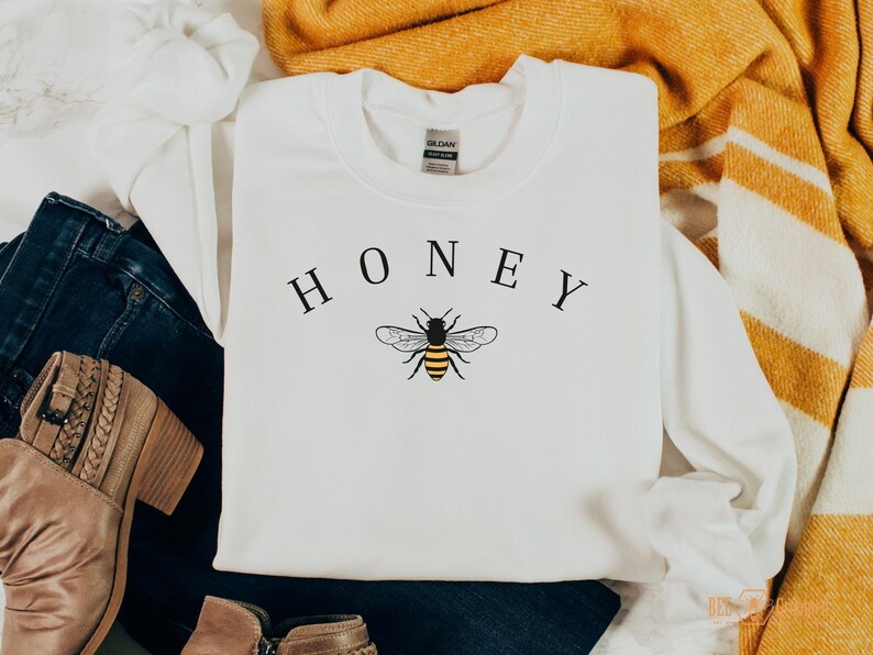 May include: White sweatshirt with the word "HONEY" printed in black letters and a black and yellow bee illustration.