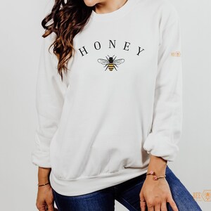 May include: White sweatshirt with the word "HONEY" printed in black letters and a small bee graphic in black and yellow.