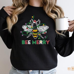 May include: Black sweatshirt with a Christmas bee design. The bee is wearing a Santa hat and is surrounded by Christmas lights. The text "BEE MERRY" is printed below the bee.