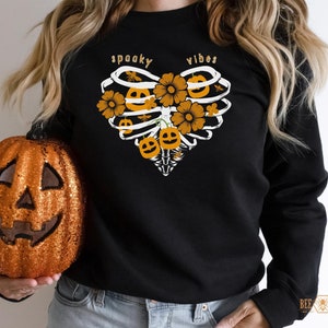 May include: Black sweatshirt with a graphic of a heart-shaped ribcage with pumpkins and flowers. The text "spooky vibes" is above the graphic.