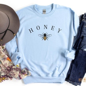 May include: Light blue sweatshirt with the word "HONEY" printed in black above a graphic of a bee. The bee is yellow and black with brown wings.