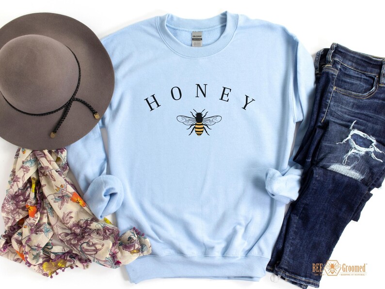 May include: Light blue sweatshirt with the word "HONEY" printed in black above a graphic of a bee. The bee is yellow and black with brown wings.