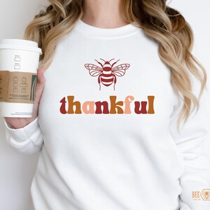 May include: White sweatshirt with a brown and orange bee graphic and the word "thankful" in brown, orange, and pink letters.