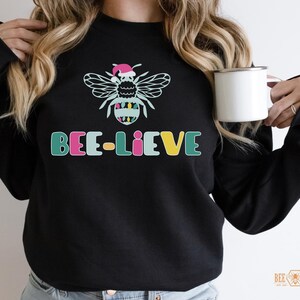 May include: A black sweatshirt with a colorful bee wearing a Santa hat and the text "BEE-LIEVE".