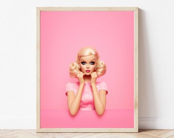 May include: A framed print featuring a doll with blonde hair and a pink dress against a pink background. The doll is posed with hands near her face. The frame is a light wood color, and the overall aesthetic is bright and playful.