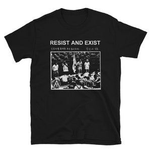 Op de afbeelding: Zwart-wit T-shirt met een grafische print met de tekst "Resist and Exist" en een foto van een groep demonstranten. De tekst in het Koreaans luidt "민주주의를 향한 싸움은 계속되어야 한다 - 핟고 나 살"