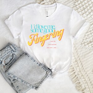 May include: White t-shirt with the text "I'd love me some good Fingering... and DK, and worsted, and aran, and..." in yellow and blue lettering.
