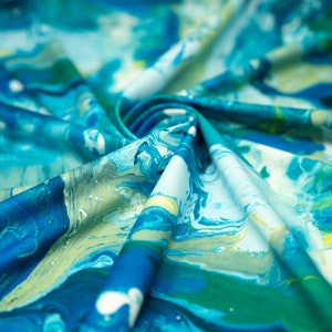 Op de afbeelding: Abstracte blauwe, groene en witte stof met een marmerpatroon. De stof is zacht en vloeibaar en creëert een gevoel van beweging.