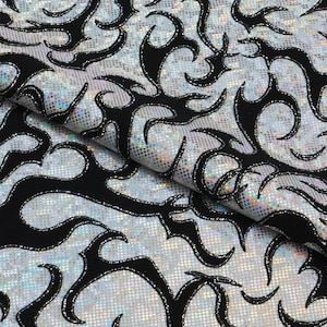 May include: A close-up of a fabric with a silver, dotted background and black flame-like designs. The fabric has a shiny, iridescent quality, with the black designs outlined in silver glitter. The fabric is folded over, showing the pattern's repeating design.