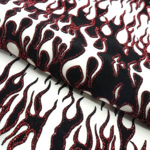 May include: Close-up of a white fabric featuring a repeating flame design. The flames are black with red glitter edging. The fabric is folded, displaying the texture and pattern.