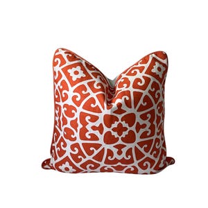 May include: Decorative throw pillow with a white and orange geometric pattern. The pillow features a repeating design of stylized floral and scroll shapes. The pillow is square and has a soft, inviting appearance, suitable for home decor.