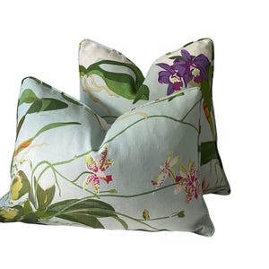 K&ouml;nnte beinhalten: Zwei dekorative Kissen mit hellblauem Hintergrund und einem floralen Muster mit Orchideen in Lila, Rosa, Gelb und Gr&uuml;n. Die Kissen haben ein weiches Aussehen und werden wahrscheinlich f&uuml;r die Inneneinrichtung verwendet.