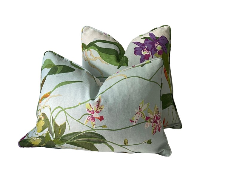 K&ouml;nnte beinhalten: Zwei dekorative Kissen mit hellblauem Hintergrund und einem floralen Muster mit Orchideen in Lila, Rosa, Gelb und Gr&uuml;n. Die Kissen haben ein weiches Aussehen und werden wahrscheinlich f&uuml;r die Inneneinrichtung verwendet.