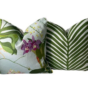 K&ouml;nnte beinhalten: Zwei dekorative Kissen. Eines hat einen hellblauen Hintergrund mit einem Blumendesign aus lila Orchideen und gr&uuml;nen Bl&auml;ttern. Das andere Kissen hat ein gr&uuml;n-wei&szlig;es Streifenmuster, das einem gro&szlig;en Blatt &auml;hnelt.