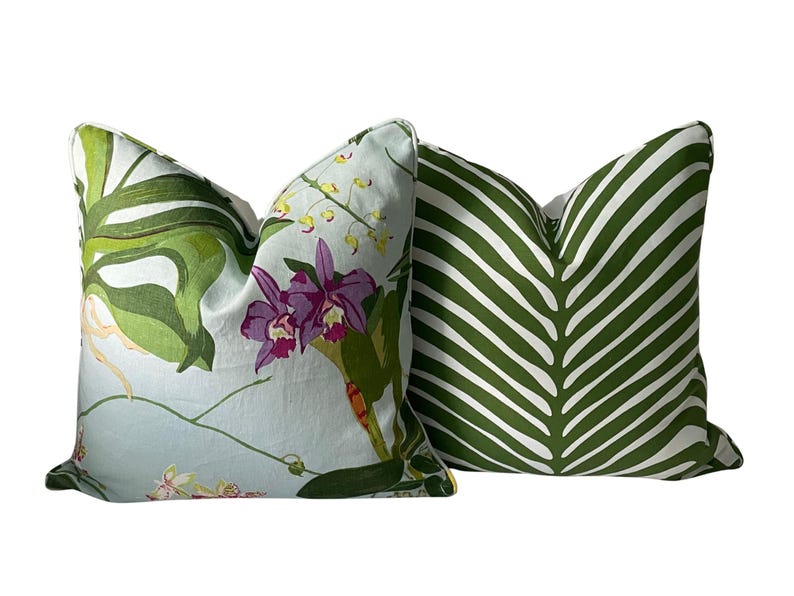 K&ouml;nnte beinhalten: Zwei dekorative Kissen. Eines hat einen hellblauen Hintergrund mit einem Blumendesign aus lila Orchideen und gr&uuml;nen Bl&auml;ttern. Das andere Kissen hat ein gr&uuml;n-wei&szlig;es Streifenmuster, das einem gro&szlig;en Blatt &auml;hnelt.
