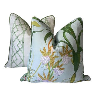 K&ouml;nnte beinhalten: Zwei dekorative Kissen. Eines zeigt ein Bambusgittermuster in Gr&uuml;n auf wei&szlig;em Hintergrund. Das andere hat einen hellblauen Hintergrund mit einem Blumendesign aus Orchideen und gr&uuml;nen Bl&auml;ttern. Beide Kissen haben gr&uuml;ne Paspeln.