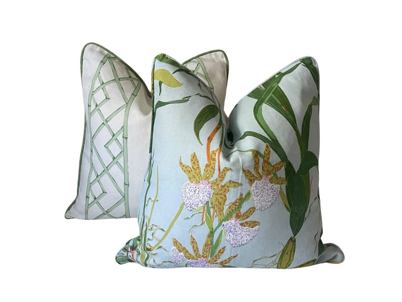 K&ouml;nnte beinhalten: Zwei dekorative Kissen. Eines zeigt ein Bambusgittermuster in Gr&uuml;n auf wei&szlig;em Hintergrund. Das andere hat einen hellblauen Hintergrund mit einem Blumendesign aus Orchideen und gr&uuml;nen Bl&auml;ttern. Beide Kissen haben gr&uuml;ne Paspeln.