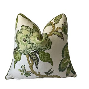 May include: A square throw pillow featuring a cream-colored fabric base with a botanical print of large green flowers and brown branches. The pillow is finished with a solid green piped edge that outlines the entire perimeter of the cushion.