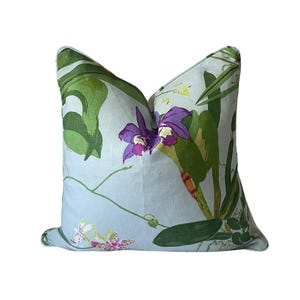 K&ouml;nnte beinhalten: Ein quadratisches Zierkissen mit hellblauem Hintergrund und floralem Muster. Das Design zeigt gro&szlig;e gr&uuml;ne Bl&auml;tter, lila Orchideen und kleinere wei&szlig;e und rosa Bl&uuml;ten. Ein dekoratives Wohnaccessoire.