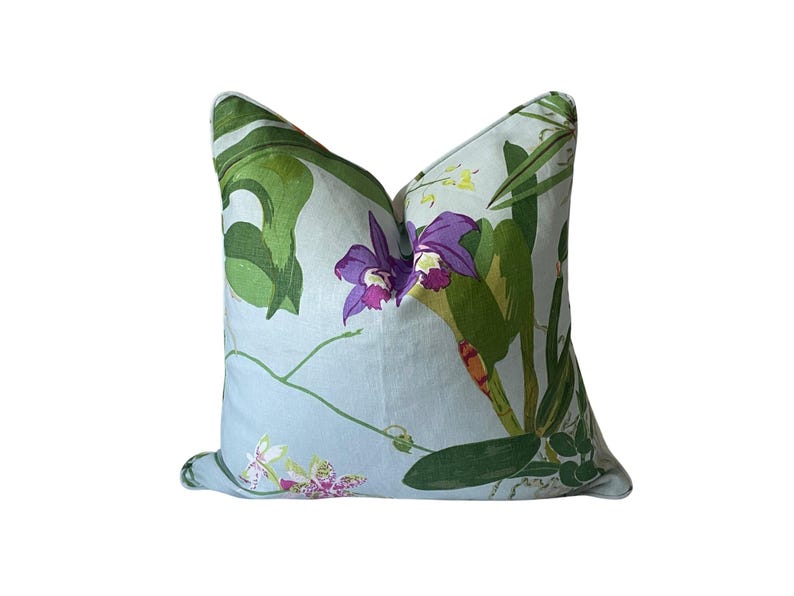 K&ouml;nnte beinhalten: Ein quadratisches Zierkissen mit hellblauem Hintergrund und floralem Muster. Das Design zeigt gro&szlig;e gr&uuml;ne Bl&auml;tter, lila Orchideen und kleinere wei&szlig;e und rosa Bl&uuml;ten. Ein dekoratives Wohnaccessoire.