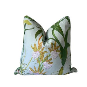 K&ouml;nnte beinhalten: Ein quadratisches Zierkissen mit hellblauem Hintergrund und einem botanischen Druck. Der Druck zeigt gr&uuml;ne Bl&auml;tter, gelbe und braune Orchideen und wei&szlig;e Akzente. Das Kissen hat ein weiches, einladendes Aussehen.