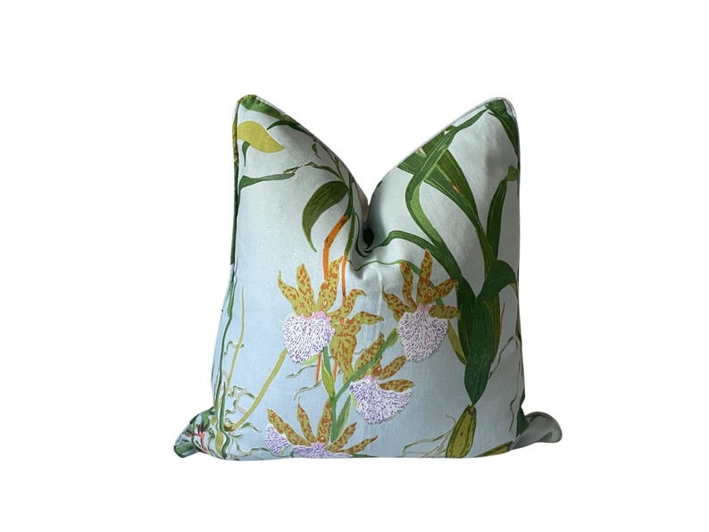 K&ouml;nnte beinhalten: Ein quadratisches Zierkissen mit hellblauem Hintergrund und einem botanischen Druck. Der Druck zeigt gr&uuml;ne Bl&auml;tter, gelbe und braune Orchideen und wei&szlig;e Akzente. Das Kissen hat ein weiches, einladendes Aussehen.