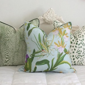 K&ouml;nnte beinhalten: Drei dekorative Kissen mit verschiedenen Mustern. Eines hat ein hellgr&uuml;nes Holzmaserungsdesign mit schwarzem Rand. Ein anderes hat ein Blumenmuster mit gr&uuml;nen Bl&auml;ttern und lila Bl&uuml;ten auf hellblauem Hintergrund. Das dritte hat einen wei&szlig;en Hintergrund mit gr&uuml;nen Punkten.
