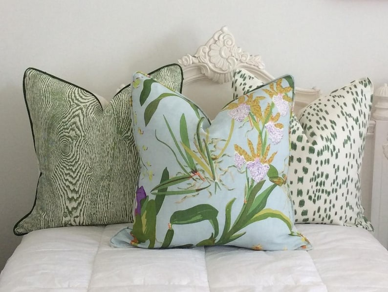 K&ouml;nnte beinhalten: Drei dekorative Kissen mit verschiedenen Mustern. Eines hat ein hellgr&uuml;nes Holzmaserungsdesign mit schwarzem Rand. Ein anderes hat ein Blumenmuster mit gr&uuml;nen Bl&auml;ttern und lila Bl&uuml;ten auf hellblauem Hintergrund. Das dritte hat einen wei&szlig;en Hintergrund mit gr&uuml;nen Punkten.