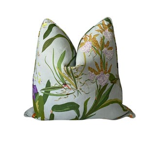 K&ouml;nnte beinhalten: Ein quadratisches Zierkissen mit floralem Muster. Der hellblaue Stoff ist mit Orchideen, gr&uuml;nen Bl&auml;ttern und lila Bl&uuml;ten bedruckt. Ein dekoratives Element, das jedem Raum einen Hauch von Natur verleiht.