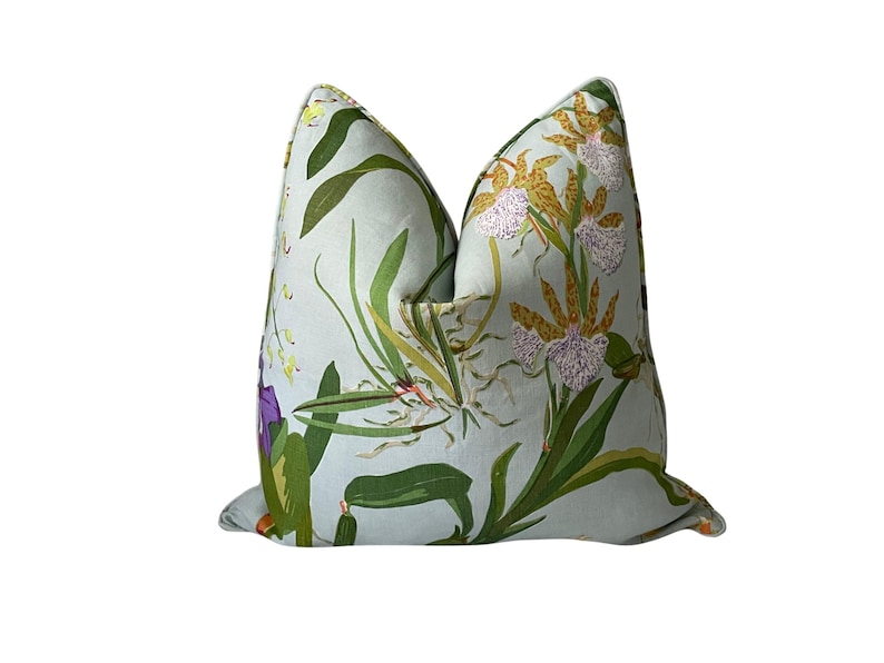 K&ouml;nnte beinhalten: Ein quadratisches Zierkissen mit floralem Muster. Der hellblaue Stoff ist mit Orchideen, gr&uuml;nen Bl&auml;ttern und lila Bl&uuml;ten bedruckt. Ein dekoratives Element, das jedem Raum einen Hauch von Natur verleiht.