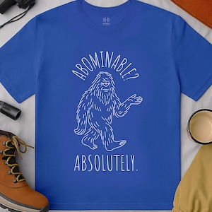 May include: Royal blue t-shirt with a white illustration of a Bigfoot and the text "ABOMINABLE? ABSOLUTELY." The shirt is surrounded by outdoor gear, including a brown boot, a black binocular, and an orange beanie.