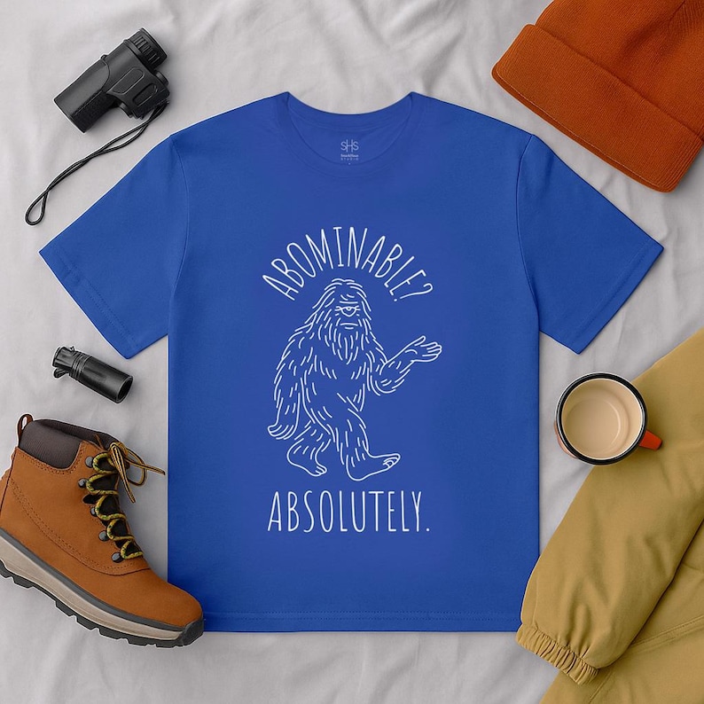 May include: Royal blue t-shirt with a white illustration of a Bigfoot and the text "ABOMINABLE? ABSOLUTELY." The shirt is surrounded by outdoor gear, including a brown boot, a black binocular, and an orange beanie.
