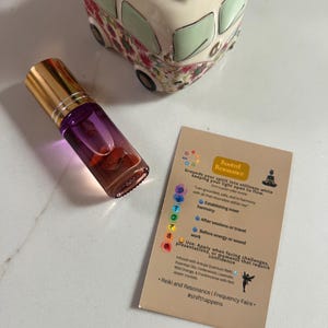May include: A purple and clear essential oil roller bottle with a gold cap, next to a card with text about Reiki and Resonance. The card has a colorful chakra chart and the words "Rooted Resonance". A decorative bus-shaped planter is in the background.