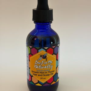 May include: A cobalt blue glass bottle with a black dropper top. The label features a bee logo and the text "Bee For Me Naturally, Growth Mint In Thyme Hair and Scalp Oil, 2 fl oz."