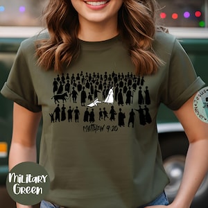 May include: Military green t-shirt featuring a black graphic of a crowd of people, a bull, and a figure in white. The text "Matthew 9:20" is below the graphic. A circular logo with text is on the right sleeve.