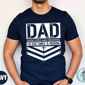 May include: Navy blue t-shirt with a white graphic that says "DAD DEDICATED AND DEVOTED TO GOD, FAMILY, & FREEDOM." The graphic also includes a white cross and chevron design.