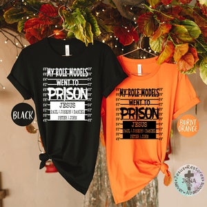 May include: Two t-shirts with a black and burnt orange color scheme. The shirts feature a white graphic with the text "My Role Models Went To Prison" and a list of names including "Jesus, Paul, Joseph, Daniel, Peter, John".