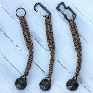 May include: Three brown and black paracord keychains with metal carabiner clips and a small, round, textured weight at the bottom. One keychain has a ring, and the other two have different styles of carabiner clips.