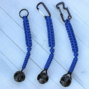 May include: Three blue paracord keychains with black metal carabiner clips and a black ball at the end. The keychains are displayed on a light blue wooden surface.