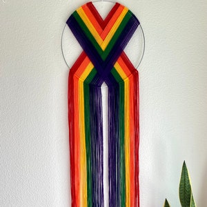 May include: A rainbow-colored wall hanging with a circular metal frame. The design features vertical strands of red, orange, yellow, green, blue, and purple yarn. The yarn is arranged in a symmetrical pattern, creating a vibrant and eye-catching decorative piece.
