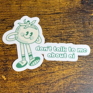 May include: A white sticker featuring a cartoon tomato character with green accents. The tomato has arms, legs, and a smiling face. Text on the sticker reads "don't talk to me about ai".