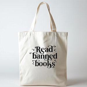 Può includere: Borsa tote color crema con la scritta "Read banned books" in corsivo nero. La borsa ha due manici ed è realizzata in un tessuto robusto. Il testo è centrato sulla parte anteriore della borsa, con accenti di stelle.