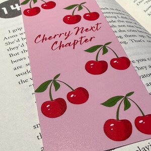 May include: A pink bookmark with a repeating pattern of red cherries and green leaves. The text "Cherry Next Chapter" is written in a cursive font. The bookmark is placed on top of an open book.